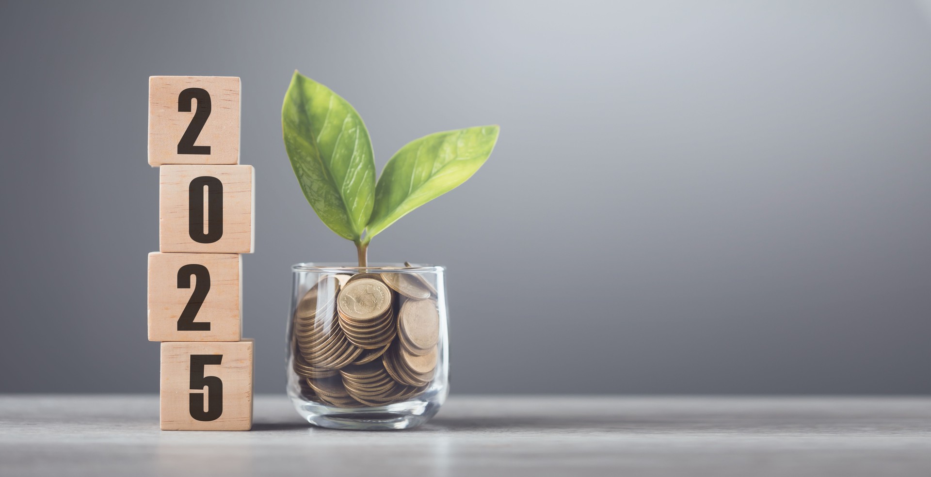 2025 is written on a wooden block next to a glass jar filled with coins, placed on a wooden table against a bright white. symbolizes financial planning, savings, and effective money management